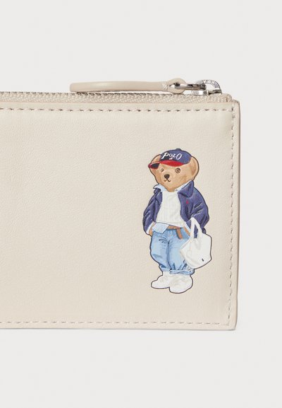 Μπεζ δερμάτινο πορτοφόλι με σχέδιο καρτούν αρκούδας σε μπλε σακάκι και λευκό πουκάμισο, κρατώντας μια λευκή τσάντα, με λεπτομερείς κεντήματα.