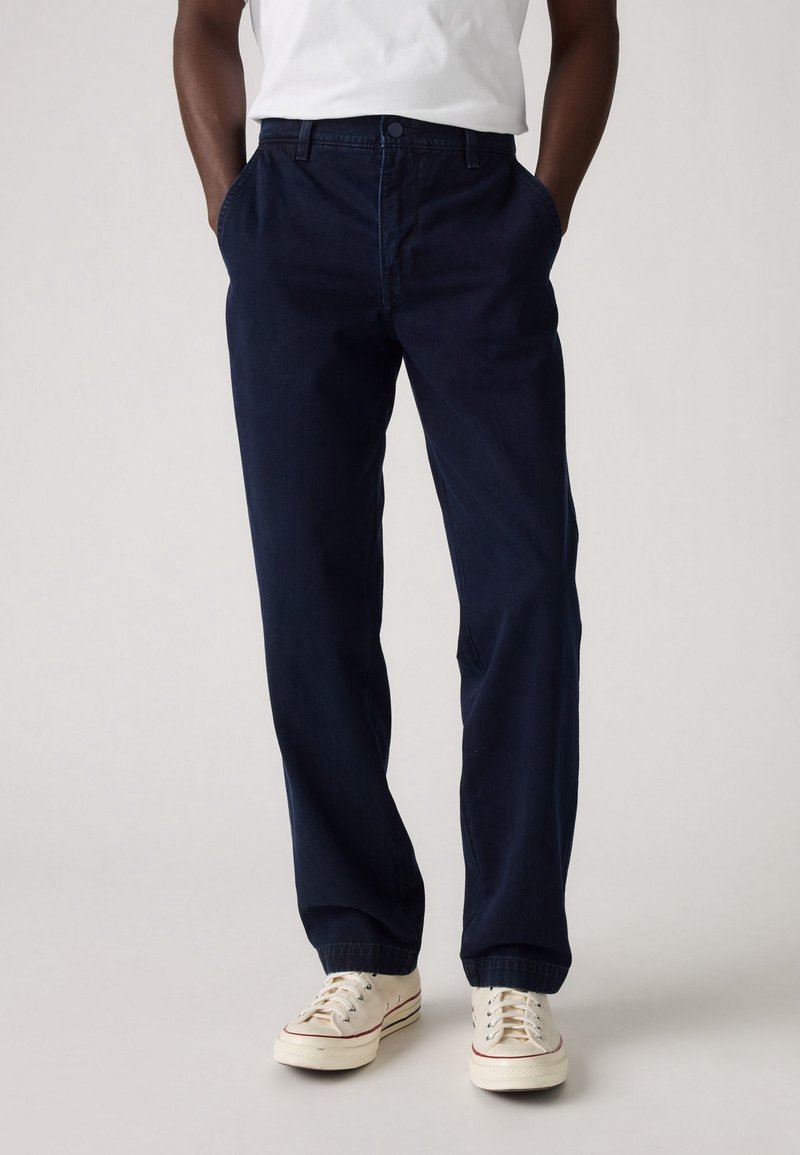 Personne portant un pantalon bleu marine foncé à coupe ample avec des poignets élastiques, associé à des baskets blanches et un t-shirt blanc, mains dans les poches.