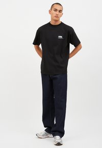 Svart bomullströja med korta ärmar och rundad hals; har en liten flerfärgad grafik på bröstet. Matchas med mörkblåjeans.