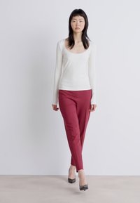 Witte lange mouwen top met een ronde hals, gecombineerd met aangesloten bordeauxrode broek. Het model draagt grijze hoge hakken. Neutrale achtergrond.