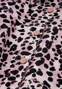 Roze stof met een zwart gevlekt patroon, voorzien van metallic knopen en uitgesproken stiksels. De textuur lijkt zacht en iets fluweelachtig.