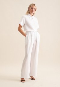 Camicia bianca di lino a maniche corte con colletto a punta, abbinata a pantaloni bianchi di lino a vita alta e gamba larga. Sandali neri con cinturini.