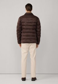 Chaqueta acolchada marrón con diseño acolchado y cuello, combinada con pantalones beige y zapatos oscuros, vista desde atrás.