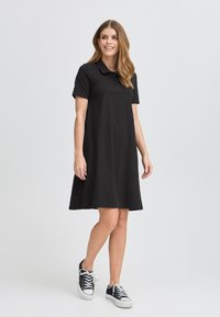 Svart polo-liknande klänning med korta ärmar, utsvängd kjol och mjuk textur; matchad med svarta sneakers. Enkel och mångsidig design.