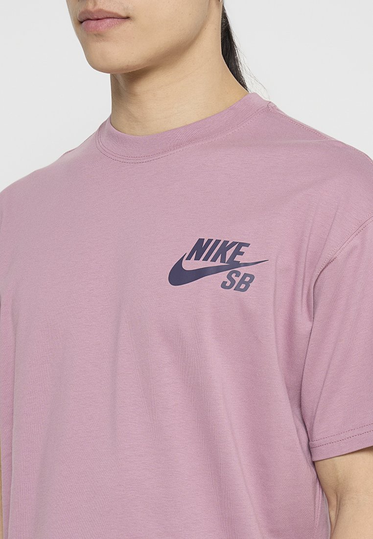 Jasnoróżowa koszulka Nike SB z krótkim rękawem, okrągłym dekoltem i ciemnoniebieskim logo nadrukowanym po lewej stronie. Miękki materiał.