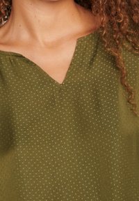 Olijfgroene blouse met een V-hals, voorzien van een stippenstructuur. De stof lijkt lichtgewicht en zacht, met een subtiele glans en stikseldetails.