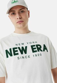 Weißes Baumwoll-T-Shirt mit grüner bestickter Aufschrift "NEW YORK NEW ERA SINCE 1920." Das Modell trägt eine hellgraue Cap mit Logo.