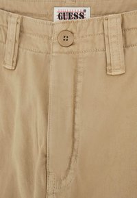 Beige katoenen broek met knoopsluiting, zichtbare stiksels en een GUESS-label op de tailleband. Gladde textuur met een klassiek ontwerp.