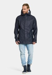 Marinblå vattentät jacka med huva, tryckknappar framtill och sidofickor. Kombinerad med ljusblå jeans och mörka stövlar. Slät textur.