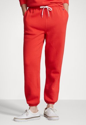 Personne portant un pantalon de jogging rouge vif avec un cordon blanc et des baskets blanches, debout les mains dans les poches devant un fond uni.
