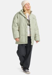 Frau in einer hellgrünen, oversized Daunenjacke, beigem Strickmütze, gemustertem Pullover, weiten schwarzen Hosen und grauen Sneakers, die nach vorne läuft.