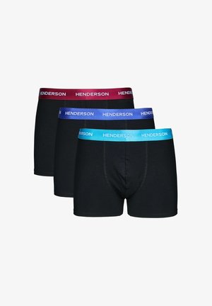 Mann mit kurzen Haaren steht oberkörperfrei und trägt schwarze Boxershorts mit einem blauen Bund, auf dem "Henderson" steht, vor einfachem Hintergrund.