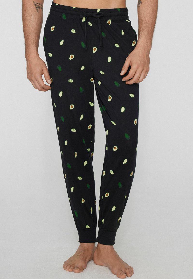 Schwarze Schlafanzughose mit grünem und beigem Avocado-Muster, elastischen Bündchen und einem Tunnelzug an der Taille, getragen von einer stehenden Person.
