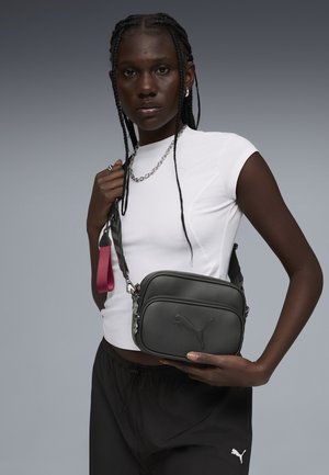 Persona de piel oscura con cabello trenzado, vistiendo una camiseta blanca y pantalones negros, sosteniendo una bolsa cruzada negra de Puma con detalle de correa rosa.