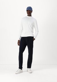 Cămașă albă cu mânecă lungă, pantaloni din corduroy bleumarin și adidași albi. Modelul poartă o șapcă albastră cu un logo. O ținută simplă și relaxată.