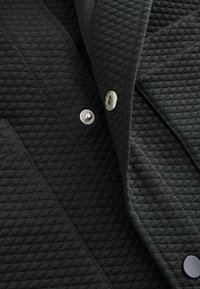 Detail einer schwarzen gesteppte Jacke, die den strukturierten Stoff, silberne Druckknöpfe und ein diagonales Nahtdesign zeigt und das Material und die Hardware betont.