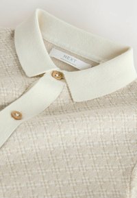 Beige knitted polo shirt with a light cream collar, wooden button closure, and textured pattern. Tag reads "NEXT, MADE IN BANGLADESH."