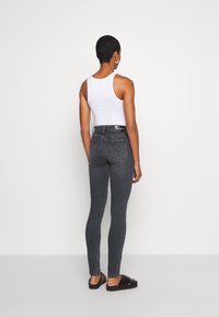 Jeans ajustados grises con una cintura alta, que presentan un diseño de bolsillo trasero y una etiqueta de cintura negra, combinados con una camiseta sin mangas blanca y sandalias negras.