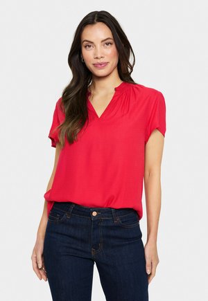 Rood korte mouwen top met een V-hals en geplooide details. De stof heeft een gestructureerd patroon, gecombineerd met donkerblauwe skinny jeans.