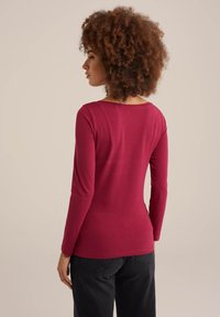 Langärmliges Oberteil in tiefem Burgunderrot, figurbetontes Design, weicher Stoff. Mit rundem Halsausschnitt und flachen Nähten, kombiniert mit dunklen Hosen.