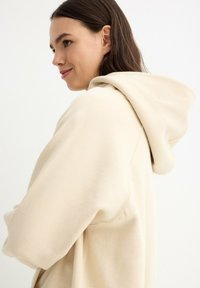 Sudadera beige con capucha y textura suave, con una capucha grande y un ajuste holgado. La tela parece de peso pesado y acogedora.