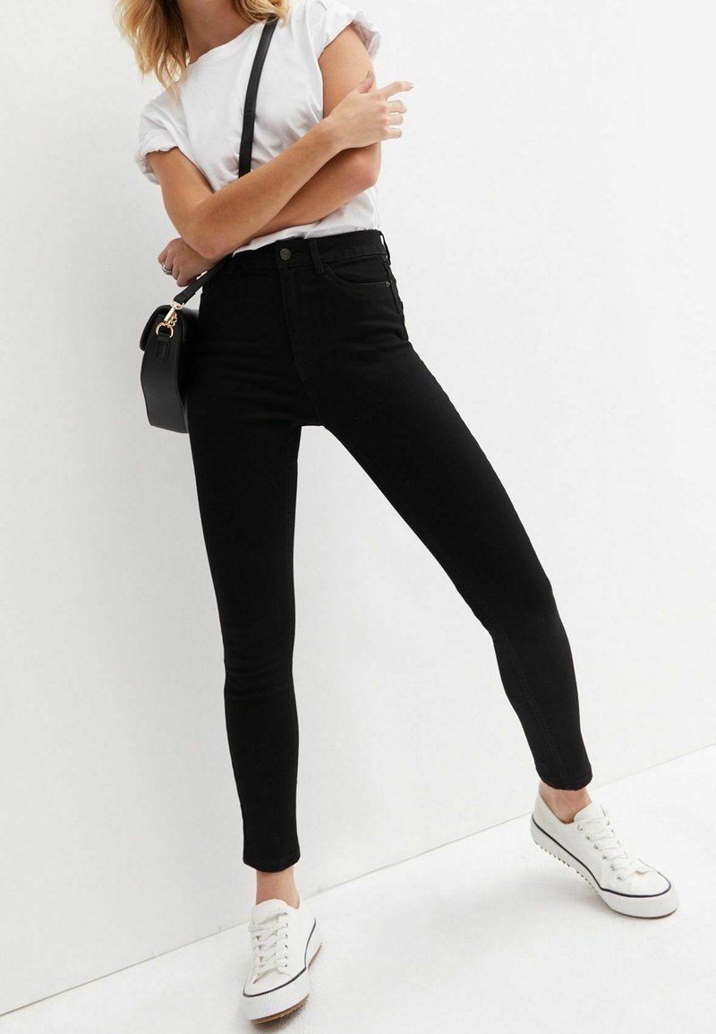 Jean skinny noir en denim, avec une taille haute, des poches avant et une texture lisse, assortis à des baskets blanches et un petit sac noir.