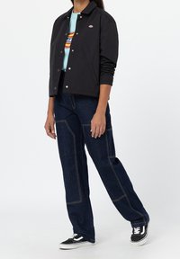 Chaqueta bomber negra, camiseta gráfica azul, vaqueros de pierna ancha en azul oscuro con costuras en contraste, y zapatillas negras con suelas blancas.