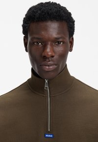 Rjava zadrga jopica z visokim ovratnikom, s srebrno zadrgo in modrim logotipom na prsih. Gladka tekstura tkanine.