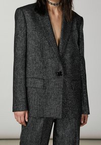 Donna che indossa un completo grigio scuro testurizzato con un unico bottone nero e pantaloni abbinati, accompagnato da una collana choker nera intrecciata.