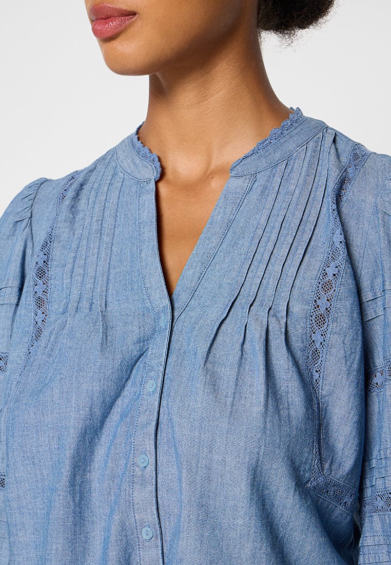 Blouse bleu clair, ressemblant à du denim, avec des manches courtes bouffantes, un devant plissé, un système de fermeture à boutons et des accents en dentelle aux épaules.