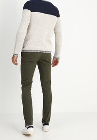 Uomo che indossa pantaloni slim-fit verde scuro, un maglione lavorato a maglia bianco e blu navy e sneakers bianche, in piedi di spalle contro uno sfondo semplice.