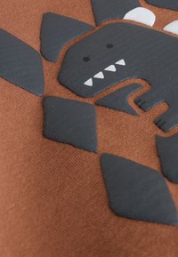 Design de monstre en caoutchouc noir sur tissu texturé couleur rouille, présentant des triangles pointus et des détails blancs sur le visage du monstre.