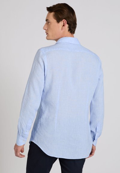 Hombre con cabello castaño vistiendo una camisa de manga larga azul claro visto desde atrás contra un fondo liso.