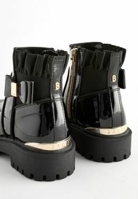 Botas de tobillo de charol negro con un cuello de textil con volantes, detalles en hardware dorado, paneles laterales elásticos y una suela negra gruesa.