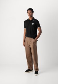 Zwart polo-shirt met witte kraagaccenten en logo, gecombineerd met beigekleurige broeken en zwarte sneakers met witte zolen. Soepele stof, ontspannen pasvorm.