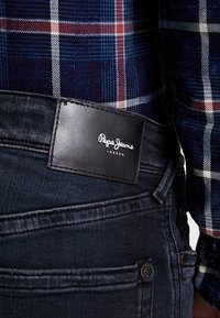 Ciemnoniebieskie dżinsy o gładkiej fakturze, z czarną skórzaną łatką z logo "Pepe Jeans London" z tyłu pasa.