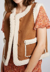 Gilet en daim marron avec un liseré en fourrure crème, doté de boutons ronds, de poches latérales et d'une finition douce et texturée. Complété par un haut floral.