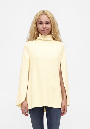 Giovane donna dai capelli biondi e ricci che indossa un maglione giallo chiaro a collo alto con maniche laterali aperte e jeans blu su uno sfondo semplice.
