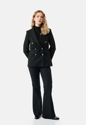 Vrouw met golvend blond haar, gekleed in een zwart dubbelgerijpt pinstripe blazer, zwarte uitlopende broek en zwarte schoenen, staand met haar handen achter de rug.