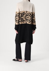 Długi kardigan z kolorowymi blokami w kremowym, beżowym i czarnym kolorze, z graficznym wzorem na środku. Dzianinowa tekstura o luźnym kroju.