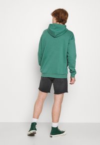 Nuori henkilö, jolla on päällään vihreä huppari, mustat farkkushortsit, valkoiset sukat ja vihreät korkeavartiset tennarit, seisoo valkoista seinää vasten.