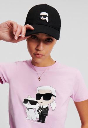Schwarze Mütze mit einem Anstecker eines Cartoon-Charakters, getragen von einer Person in einem pinken T-Shirt mit einem Grafikdesign von zwei Charakteren, von denen einer eine Katze ist.
