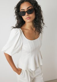 Witte peplumtop met puffed sleeves, gemaakt van zachte stof. Kenmerkt zich door een vierkante halslijn en verticale naden. Gecombineerd met crèmekleurige broeken.