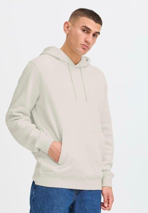 Young man wearing a light beige hoodie with a front pocket, looking confidently at the camera against a plain white background.
