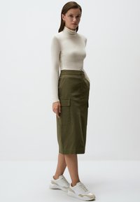 Lange mouw, crèmekleurige coltrui, gecombineerd met een olijfgroene midi-rok met cargozakken, gedragen met witte sneakers. Eenvoudige achtergrond.