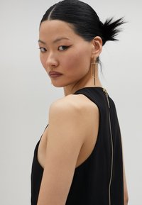 Čierne bezramienkové šaty s nízkym zakrytím chrbta a viditeľným zlatým zipsom. Modelka má na sebe dlhé geometrické zlaté náušnice; hladká pleť a tmavé vlasy.