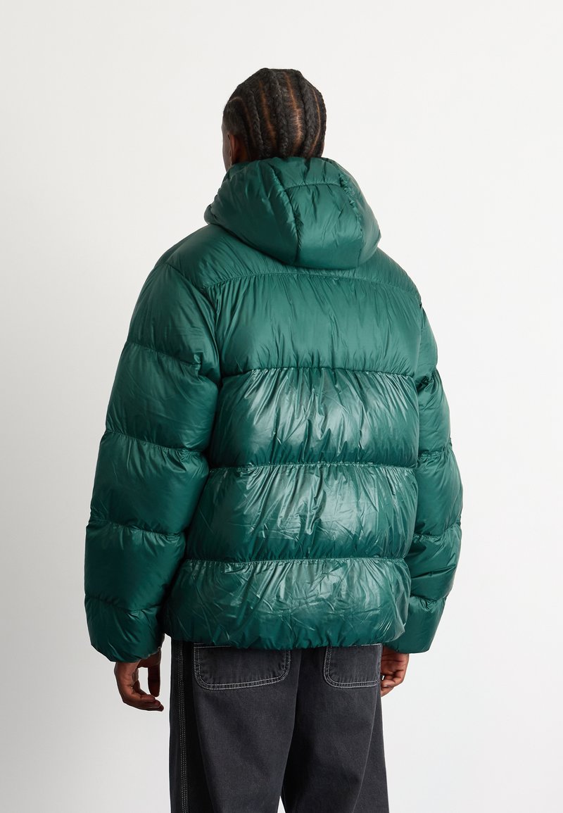 Puchowa zielona kurtka z kapturem, o błyszczącej fakturze, pikowanym wzorze i elastycznym dołem. Noszona z ciemnoszarymi spodniami.