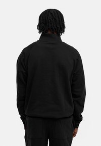 Black pullover hoodie with a high collar, long sleeves, and raglan shoulder seams. Made from textured fabric with a relaxed fit.