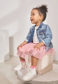 Denim jacket, white shirt, pink tulle skirt, pink socks, and white high-top sneakers. Sitting on a light circular pedestal.