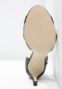 Black high-heeled shoe with a textured upper and metallic accent. Smooth, beige outsole with a rubberized grip and slender heel design.